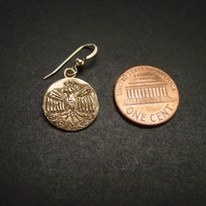 May include: A gold-colored earring with a circular pendant featuring a detailed bird design. The earring is next to a penny coin for size comparison. The coin reads "UNITED STATES OF AMERICA" and "ONE CENT".