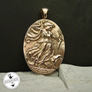 May include: Oval-shaped bronze pendant featuring a detailed relief of a figure with a bow and arrow, accompanied by a dog. The pendant has a loop at the top for a chain. The word "Handmade" is in a circle at the bottom.