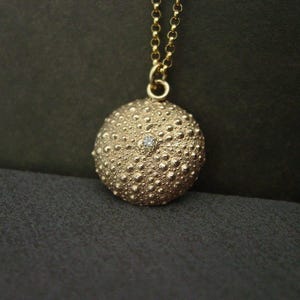 May include: A gold pendant necklace with a textured, spherical charm and a small gemstone. The charm hangs from a fine gold chain. The pendant's design is reminiscent of a sea urchin, offering a unique and stylish accessory.