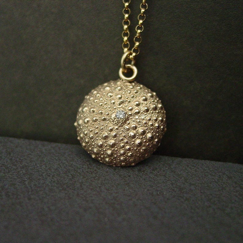 May include: A gold pendant necklace with a textured, spherical charm and a small gemstone. The charm hangs from a fine gold chain. The pendant's design is reminiscent of a sea urchin, offering a unique and stylish accessory.