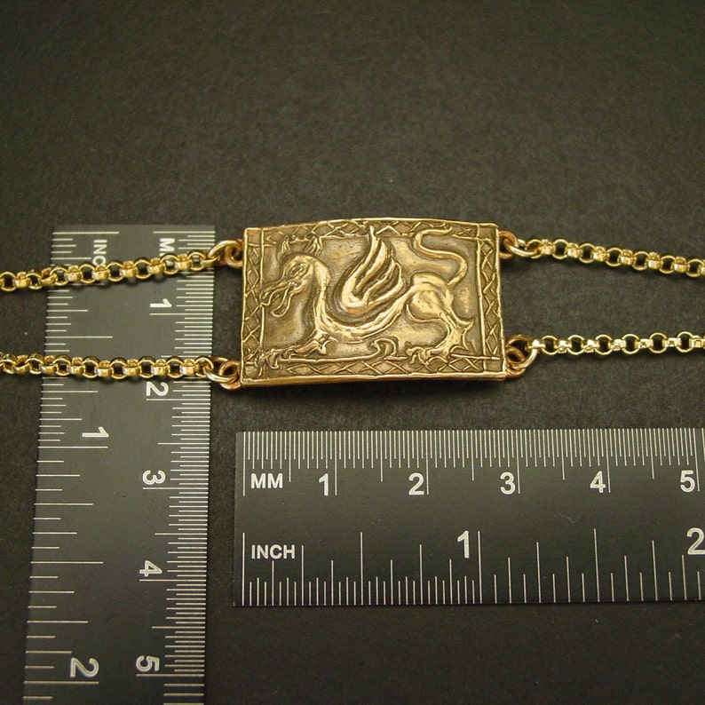 May include: A gold-toned necklace with a rectangular pendant featuring a detailed embossed design of a winged dragon. The pendant measures approximately 1 inch in length and is attached to a delicate gold chain.