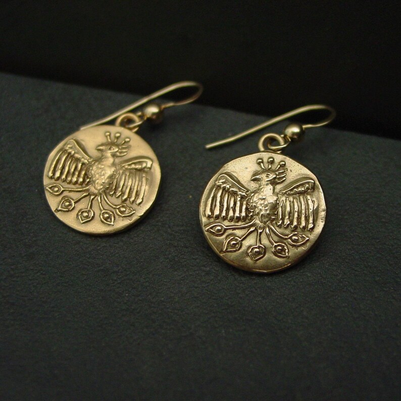 May include: A pair of gold-colored earrings. Each earring features a circular pendant with a detailed relief of a bird with outstretched wings and a crown. The earrings have a hook closure and are displayed against a dark background.