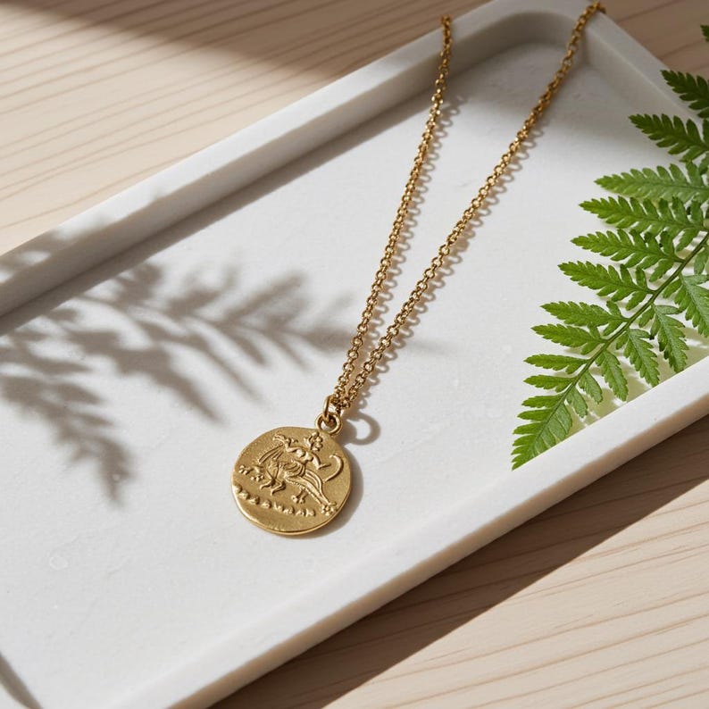 May include: A gold-toned metal necklace featuring a circular pendant with an embossed mythological figure. The pendant hangs from a delicate cable chain, showcasing a textured, coin-like design with intricate details on the surface of the metal.