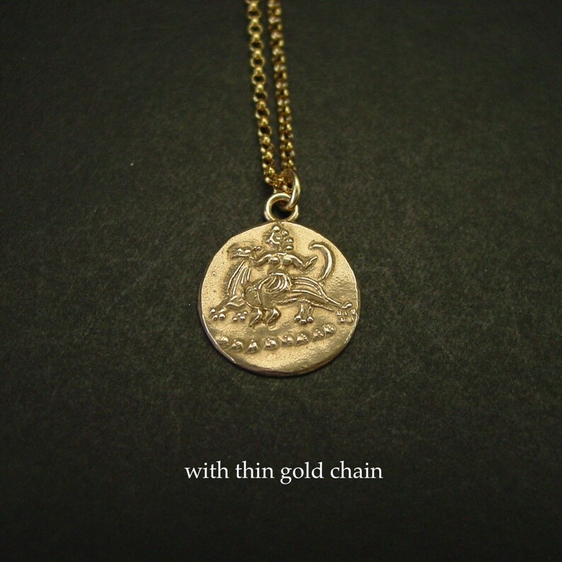 May include: A gold-toned circular pendant featuring an embossed mythological figure riding a creature, suspended from a thin gold-toned chain. The pendant displays a textured surface with the text "with thin gold chain" visible below.