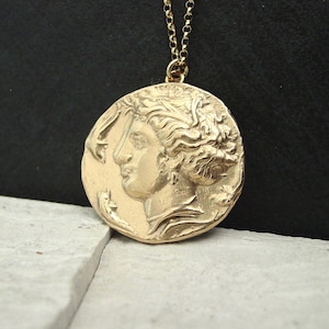 A gold round coin pendant hanging from a thin gold chain. The coin is unevenly round and has a raised profile of a woman goddess with an updo of curly hair. She is wearing chandelier earrings and a pearl necklace and is facing two dolphins.