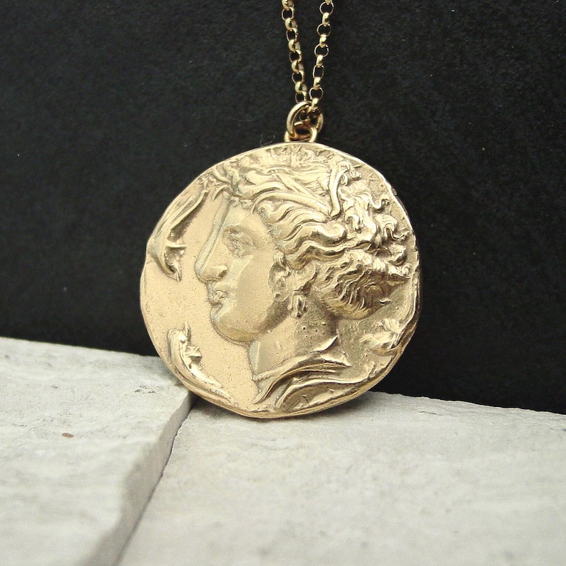 A gold round coin pendant hanging from a thin gold chain. The coin is unevenly round and has a raised profile of a woman goddess with an updo of curly hair. She is wearing chandelier earrings and a pearl necklace and is facing two dolphins.