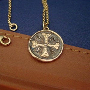 May include: A gold-toned necklace featuring a circular pendant with a textured cross design. The pendant has a dark, oxidised background within the recessed areas, contrasting with the raised, polished gold-coloured metal of the cross and border.