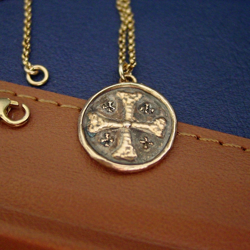 May include: A gold-toned necklace featuring a circular pendant with a textured cross design. The pendant has a dark, oxidised background within the recessed areas, contrasting with the raised, polished gold-coloured metal of the cross and border.