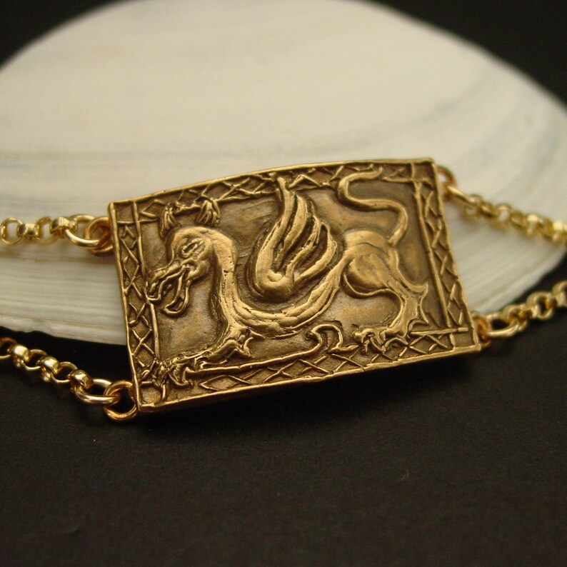 May include: A gold-toned bracelet with a rectangular pendant featuring a detailed embossed image of a winged mythical creature. The pendant is surrounded by a decorative border.