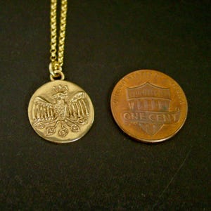 May include: A gold-toned circular pendant necklace featuring an embossed phoenix design, suspended from a delicate chain. The pendant is positioned next to a copper-colored United States one-cent coin for scale, showing a similar diameter.