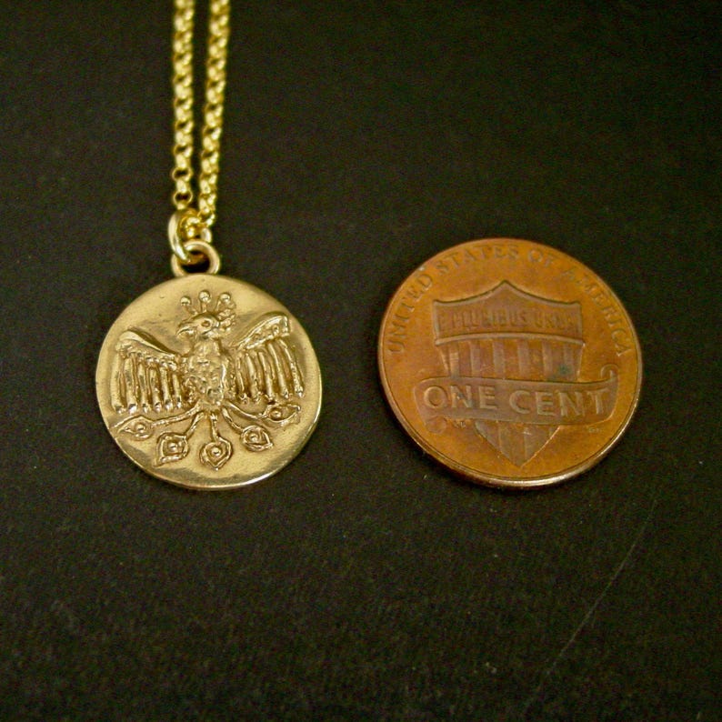 May include: A gold-toned circular pendant necklace featuring an embossed phoenix design, suspended from a delicate chain. The pendant is positioned next to a copper-colored United States one-cent coin for scale, showing a similar diameter.