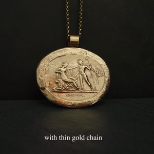 May include: Gold-toned pendant necklace with a detailed relief of classical figures. The round pendant features a seated woman and a standing man, with the text "KPEIROS" below. The necklace has a thin gold chain.