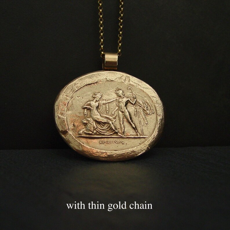 May include: Gold-toned pendant necklace with a detailed relief of classical figures. The round pendant features a seated woman and a standing man, with the text "KPEIROS" below. The necklace has a thin gold chain.