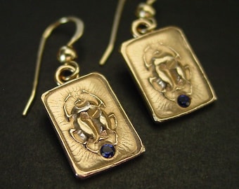 May include: Gold-toned rectangular earrings with a detailed scarab beetle design. Each earring features a small, round, blue gemstone. The earrings have a hook closure and are displayed against a dark background.