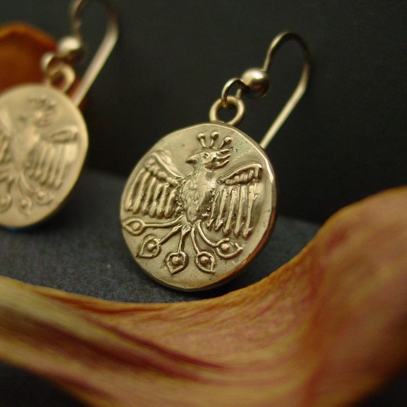 May include: Gold-toned earrings featuring a circular pendant with a detailed bird design. The bird has outstretched wings and a crown, with decorative elements below. The earrings have a simple hook closure. The background is dark.