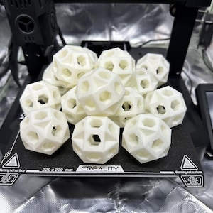 Puede incluir: Un conjunto de esferas blancas, geométricas, impresas en 3D con aberturas hexagonales. Las esferas están apiladas sobre una superficie negra con el texto "CREALITY" y "220 x 220 mm". La imagen se toma en un recinto plateado, sugiriendo una configuración de impresión 3D.