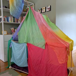 K&ouml;nnte beinhalten: Ein farbenfrohes Spielzelt, das aus Holzd&uuml;beln konstruiert und mit leuchtendem, durchsichtigem Stoff in Rot-, Rosa-, Gr&uuml;n-, Blau- und Gelbt&ouml;nen drapiert ist. Eine Regenbogenfahne h&auml;ngt von oben und erg&auml;nzt das verspielte Design.