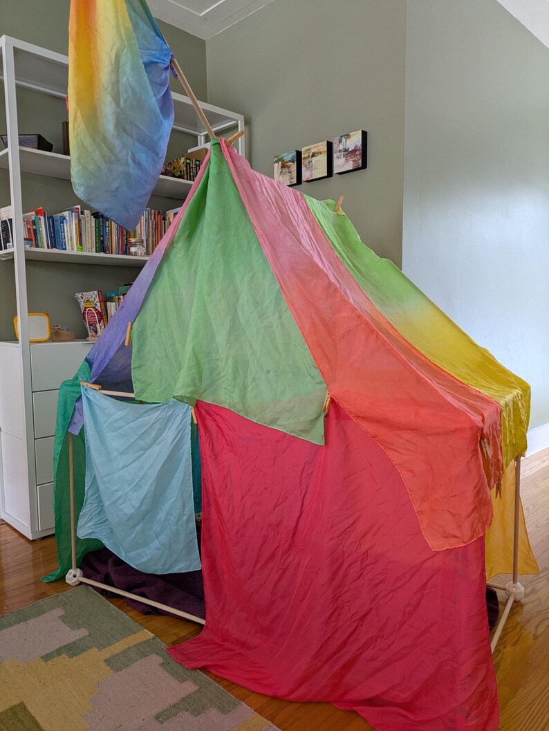 K&ouml;nnte beinhalten: Ein farbenfrohes Spielzelt, das aus Holzd&uuml;beln konstruiert und mit leuchtendem, durchsichtigem Stoff in Rot-, Rosa-, Gr&uuml;n-, Blau- und Gelbt&ouml;nen drapiert ist. Eine Regenbogenfahne h&auml;ngt von oben und erg&auml;nzt das verspielte Design.
