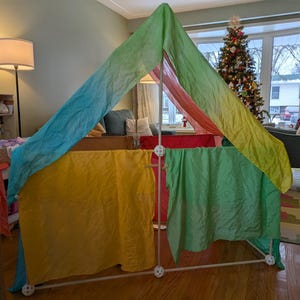 K&ouml;nnte beinhalten: Ein farbenfrohes Spielzelt mit wei&szlig;em Rahmen und Stoffbahnen in Gelb, Gr&uuml;n, Rot und Blau. Das Zelt ist in Innenr&auml;umen aufgebaut, mit einem Weihnachtsbaum im Hintergrund.