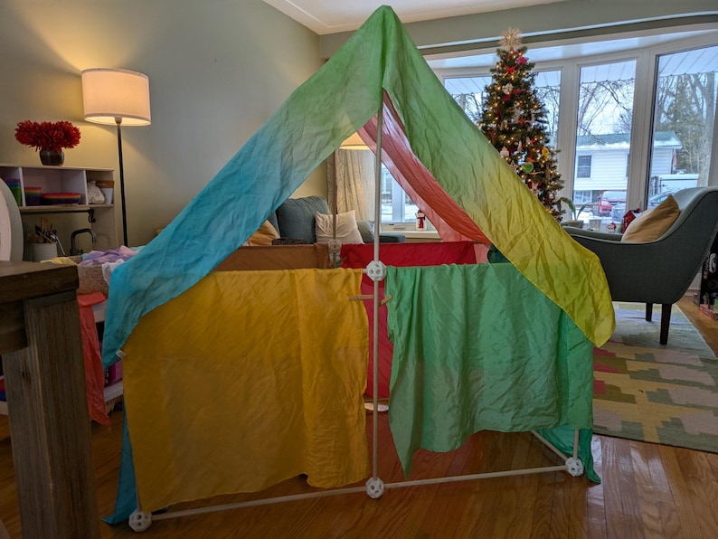 K&ouml;nnte beinhalten: Ein farbenfrohes Spielzelt mit wei&szlig;em Rahmen und Stoffbahnen in Gelb, Gr&uuml;n, Rot und Blau. Das Zelt ist in Innenr&auml;umen aufgebaut, mit einem Weihnachtsbaum im Hintergrund.