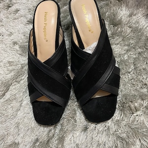 May include: A pair of black suede heeled sandals with criss-cross straps and the brand name "Hush Puppies" printed on the inside of each shoe.