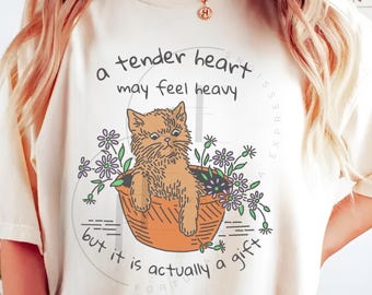May include: Ivory-colored t-shirt featuring a cartoon cat in a basket surrounded by flowers. The text reads "a tender heart may feel heavy but it is actually a gift." The shirt has a crew neck and short sleeves.