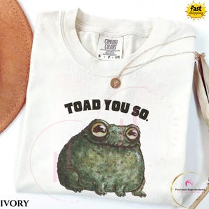 May include: Ivory t-shirt featuring the text "TOAD YOU SO." and a detailed illustration of a green toad. The shirt has a crew neck and short sleeves. A gold necklace with a pendant is draped across the shirt.