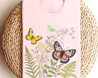 May include: A light pink t-shirt featuring a colorful butterfly and floral design. The design includes a yellow butterfly, a pink and blue butterfly, green ferns, and small white flowers. The shirt is folded on a woven, light brown surface.