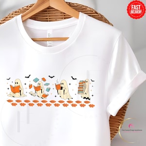 Op de afbeelding: Wit T-shirt met een Halloween-thema. De afbeelding toont stripgeesten die boeken lezen, met vleermuizen en herfstbladeren. De geesten zijn in verschillende poses, sommigen lezen, anderen houden stapels boeken vast.