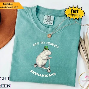 以下が含まれることがあります： 緑色の快適な色のTシャツ。緑色の帽子をかぶってスクーターに乗っているカピバラのグラフィック。テキストは「OFF TO COMMIT SHENANIGANS」と書かれています。このシャツはクルーネックと半袖です。