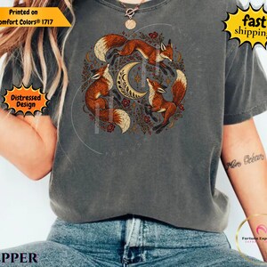 Puede incluir: Una camiseta de manga corta gris carbón con un estampado gráfico circular de tres zorros naranjas rodeando una luna creciente, acentuado con pequeñas estrellas y elementos florales. El diseño está centrado en el pecho de la prenda.