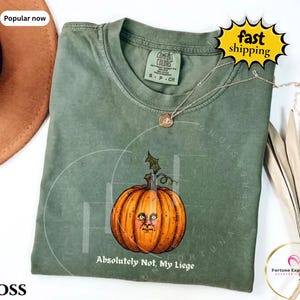 May include: A moss green short-sleeve t-shirt featuring a graphic of an orange pumpkin with a human-like face. Below the pumpkin, the text reads "Absolutely Not, My Liege". The shirt has a crew neckline and a Comfort Colors brand label inside.