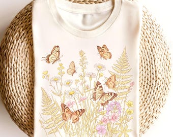 May include: Cream-colored t-shirt featuring a detailed illustration of butterflies and wildflowers. The design includes various butterflies in shades of brown and orange, amidst daisies, ferns, and other floral elements in soft pastel colors.