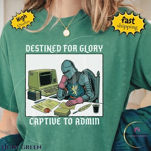 Op de afbeelding: Een bosgroen t-shirt met een afbeelding van een middeleeuwse ridder in harnas aan een bureau met een vintage computer, een stapel diskettes, een document en een ganzenveer. De tekst luidt DESTINED FOR GLORY CAPTIVE TO ADMIN.