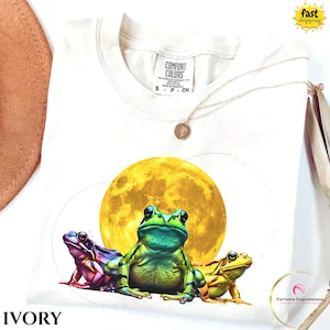 Può includere: T-shirt avorio Comfort Colors con una colorata illustrazione di tre rane davanti a una grande luna gialla. Le rane sono verdi, viola e gialle. Una collana d'oro è drappeggiata sulla maglietta.