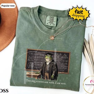 May include: A moss green crew neck t-shirt featuring a graphic of a frog wearing a graduation cap and robe standing before a chalkboard covered in mathematical equations. The text below the frog reads, The equations are fine. I am not.
