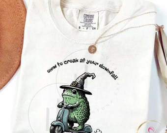 May include: Ivory-colored t-shirt with a cartoon frog wearing a witch hat and riding a scooter. The text "omw to croak at your downfall" is printed above the frog. The shirt has a Comfort Colors tag.