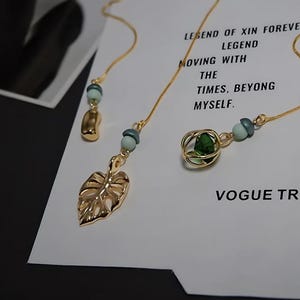 Może przedstawiać: Złote kolczyki z liściastymi i geometrycznymi zawieszkami, zaakcentowane turkusowymi koralikami. Kolczyki wiszą na delikatnych łańcuszkach. Tło zawiera tekst "Fashionable! Hand made" i "VOGUE TREND."