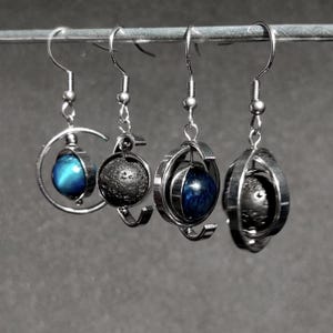 Op de afbeelding: Vier paar zilverkleurige oorbellen met unieke ontwerpen. Twee paar hebben een donkergrijze, getextureerde bol, terwijl de andere twee blauwe, bolvormige edelstenen hebben. Elke oorbel hangt aan een eenvoudige haak.