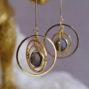 Op de afbeelding: Een paar goudkleurige oorringen. Elke oorring heeft een grijze stenen bol die is opgehangen in concentrische gouden ringen. De oorbellen zijn ontworpen met een delicate, bungelende stijl, geschikt voor dagelijks gebruik of speciale gelegenheden.