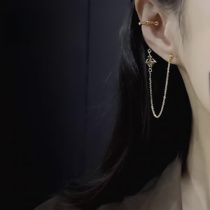 May include: Gold-coloured ear cuff and earring set. The ear cuff wraps around the upper ear, while the earring features a geometric charm and a delicate chain that connects to a stud earring. The jewellery is shown against a dark background.