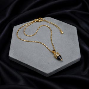 May include: A gold-toned chain necklace featuring a pendant with a pointed black stone set in a gold cap encrusted with small clear crystals. The necklace includes a delicate link chain with an adjustable clasp mechanism at the end.