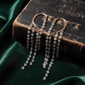 Pode incluir: Punhos de orelha dourados com um design em forma de coração, adornados com contas de cristal transparente penduradas. A joia repousa sobre um livro antigo e escuro contra um fundo de tecido de veludo verde escuro, criando uma estética luxuosa.