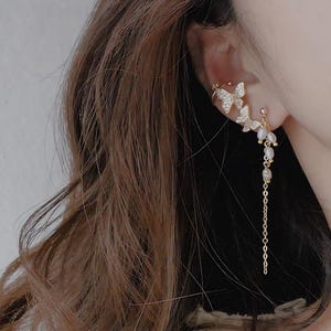 May include: Gold-toned butterfly ear cuff and dangling earring set. The ear cuff features two butterfly designs with clear crystals. The dangling earring has a chain with small pearls. The jewelry is worn on the ear.