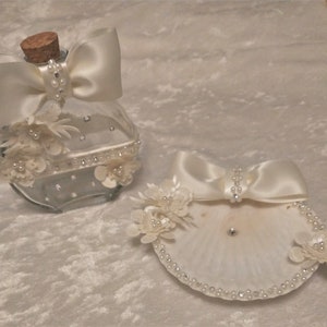 May include: Two white decorative containers for wedding rings. One is a glass bottle with a white satin bow and pearl embellishments. The other is a white seashell with a white satin bow and pearl embellishments.