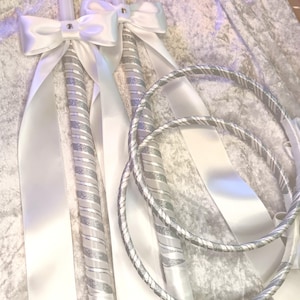 May include: Wedding ceremony set featuring two white candles with silver glitter stripes and white satin bows. Two matching silver and white rope-style rings are also included. The set is designed for a wedding ceremony.