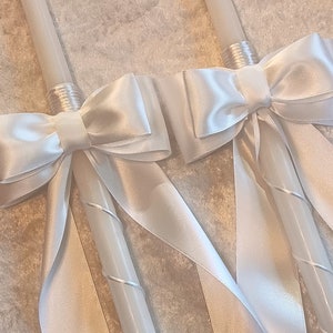 May include: Two white candles adorned with large, elegant white satin bows. The bows are centered on the candles and have long, flowing ribbons. The candles have a spiral design and are likely for a special occasion.