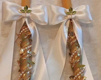 May include: Two white satin ribbon-wrapped candles with gold and green accents. The candles are decorated with gold glitter, green leaves, and white pearls.