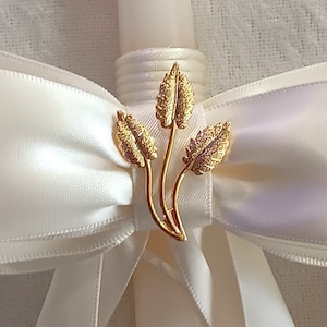 Può includere: Un fiocco di raso bianco con una spilla a forma di foglia d'oro. Il fiocco è legato attorno a una candela bianca.