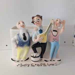 May include: A vintage ceramic planter featuring three singing figures in colorful attire. The figures are posed around a rectangular vase, with the words "Jolly Good Fellows" written on the base. The figures are wearing suits and playing a musical instrument.